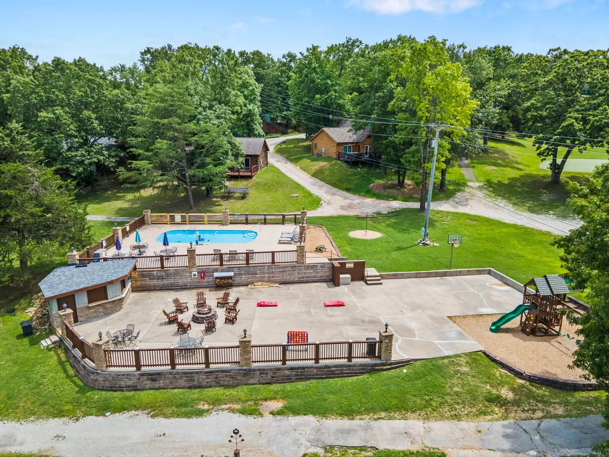 This image features a vibrant swimming pool area with a patio, nearby playground, and a basketball hoop, all surrounded by green trees. The scene includes families and children enjoying the amenities, with options for swimming, sunbathing, and children's play.