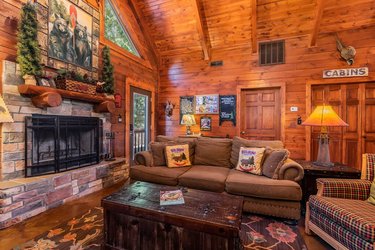 A cozy cabin interior with a stone fireplace, plush sofa, warm wooden beams, and playful bear-themed artwork. The room exudes warmth and a private, welcoming atmosphere perfect for relaxing after a day of activities.