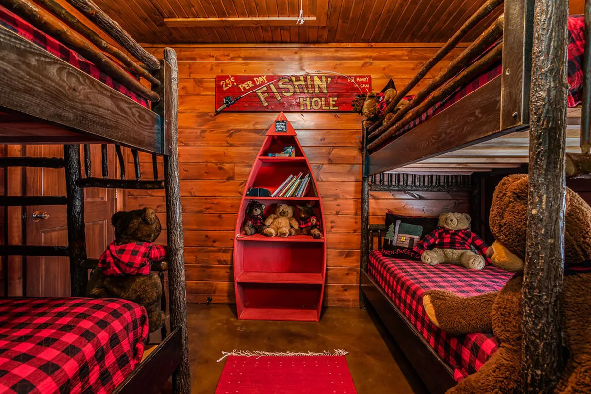 A fun and whimsical bunk room featuring two sets of twin bunks with red and black plaid bedding, playful decor like a fishing hole sign, a red boat-shaped shelf, and stuffed animals. The space is designed for kids and teens, offering a playful retreat with cozy bedding and creative touches.