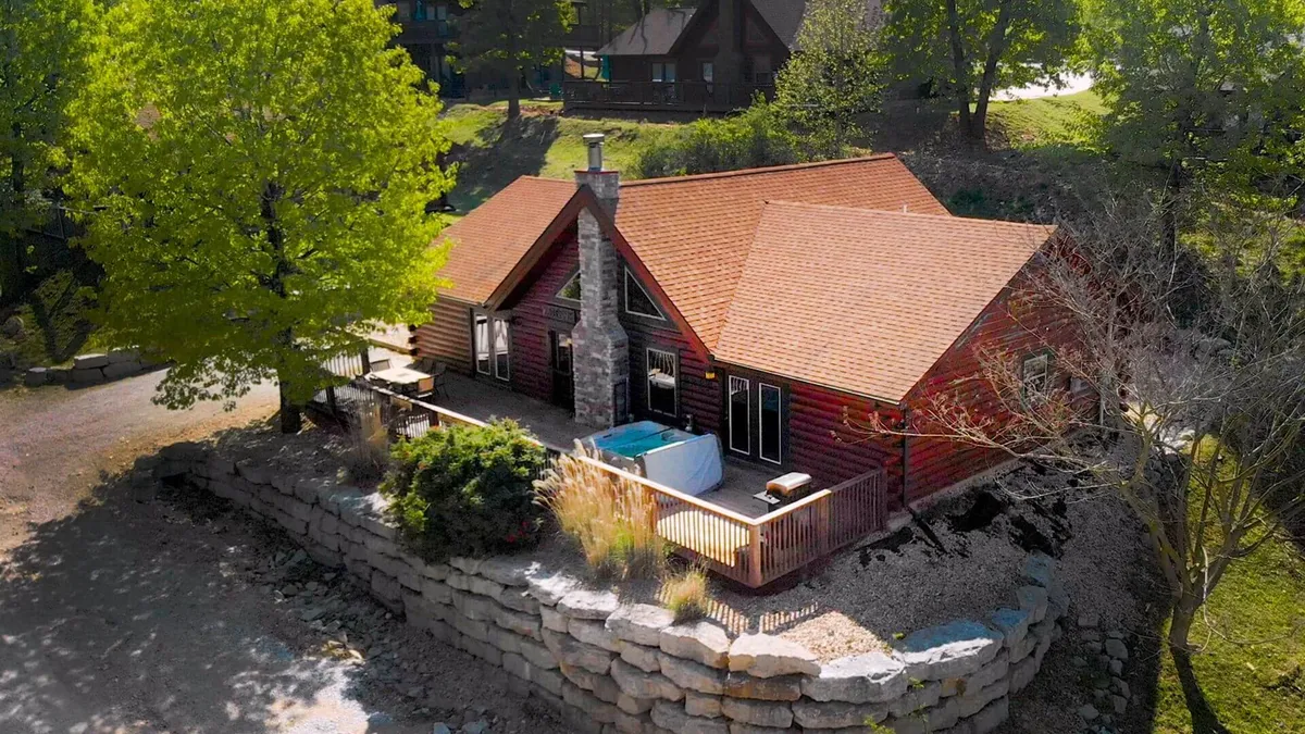 The image features a beautiful, custom-decorated real log cabin with a striking stone chimney, a spacious deck outfitted with a private hot tub, and lush greenery all around. The deck invites guests to relax, dine outdoors, or enjoy the serenity of the natural setting, with opportunities for stargazing or cozy evening gatherings by the fire pit.