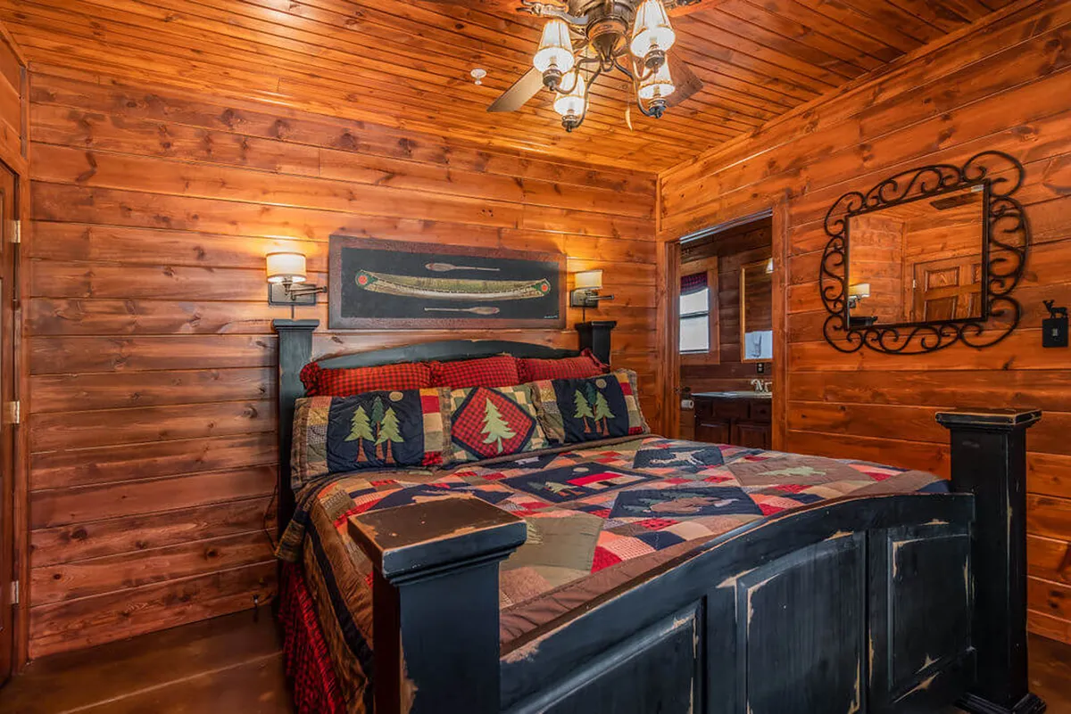 This image showcases a king bedroom with a wooden bed frame, a colorful quilt, wooden walls, chandelier, and decorative mirror. The room exudes a warm, inviting atmosphere perfect for restful nights. The bed is large and plush, with soft lighting and rustic decor highlighting comfort and charm.