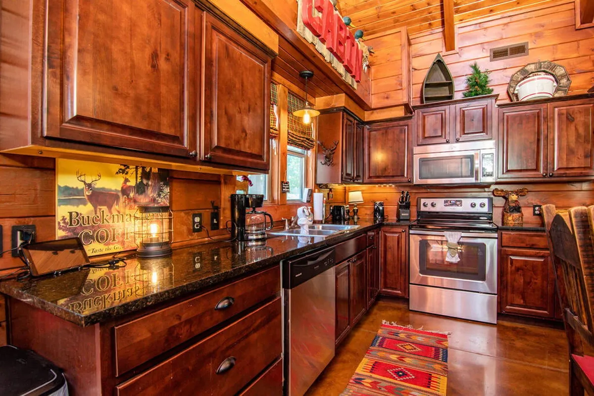 A close-up of a rustic kitchen featuring dark wooden cabinetry, granite countertops, a colorful rug, and modern kitchen appliances. The setup feels both welcoming and functional, inviting guests to cook and socialize.