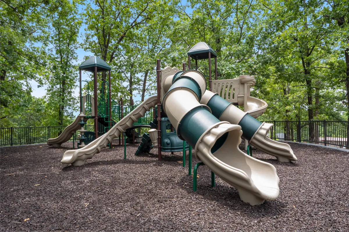 A vibrant playground area features slides, climbing structures, and is surrounded by tall green trees. Children and families can be seen playing and socializing, highlighting the fun and safe environment for younger guests.