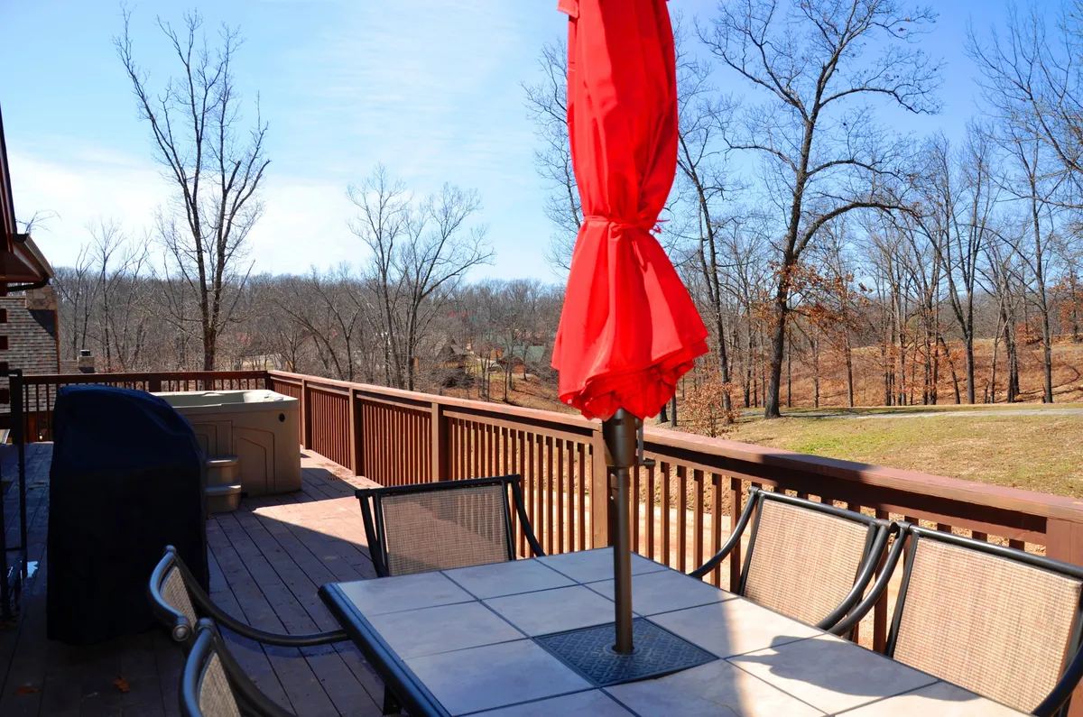 A cozy outdoor dining setup includes a red umbrella shading a dining table and a propane grill nearby, all set against a backdrop of trees. The arrangement suggests ease of outdoor meals, barbecues, and gatherings in a forested environment.
