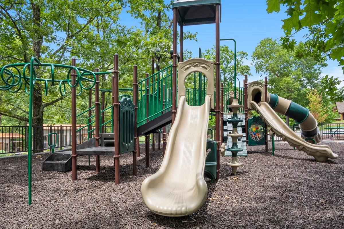 This image shows a colorful playground with slides and climbing structures among tall trees. Children are seen playing and socializing, while the lush natural backdrop emphasizes the outdoor, family-friendly setting.