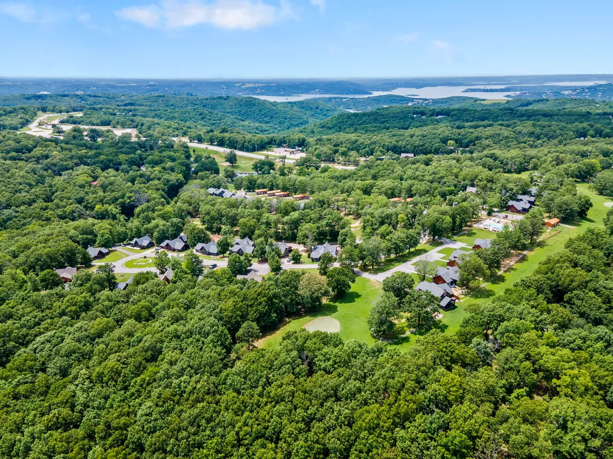 An expansive view over a lush, green forest, with the neighborhood layout visible in the foreground and a shimmering lake in the distance. The scene conveys a sense of scale, adventure, and access to diverse outdoor activities like hiking and golfing.