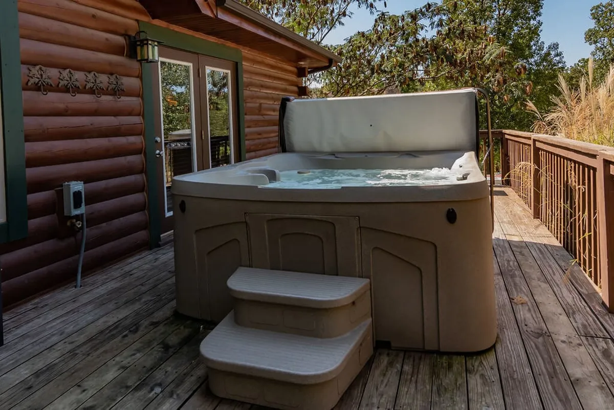 A serene wooden deck attached to a log cabin features a bubbling hot tub with a view of lush greenery. The spacious deck also includes ample seating and is situated in a tranquil, natural setting, making it ideal for unwinding and connecting with nature.