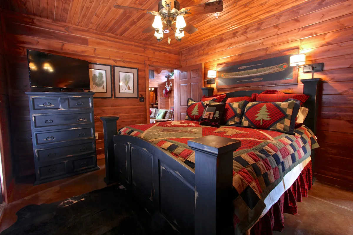 A king bedroom with a decorative quilt on the bed, a chest of drawers, wall-mounted TV, and artwork. The room feels warm and welcoming, with plush bedding and thoughtful decor for relaxation and entertainment.