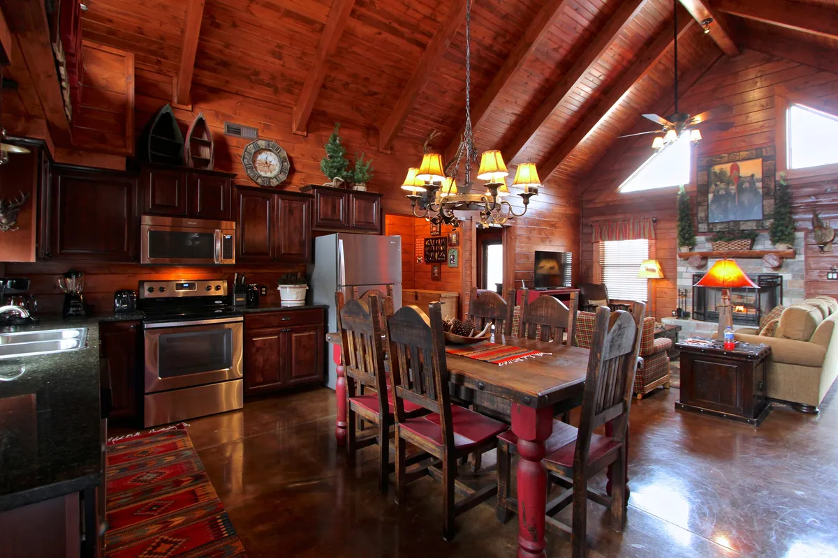 A wide view of an open-plan great room, featuring a plush sofa near a stone fireplace, a large wooden dining table beneath a rustic chandelier, and a well-equipped kitchen with modern appliances. The space is bathed in natural light, with clear sightlines connecting the living, dining, and kitchen areas.